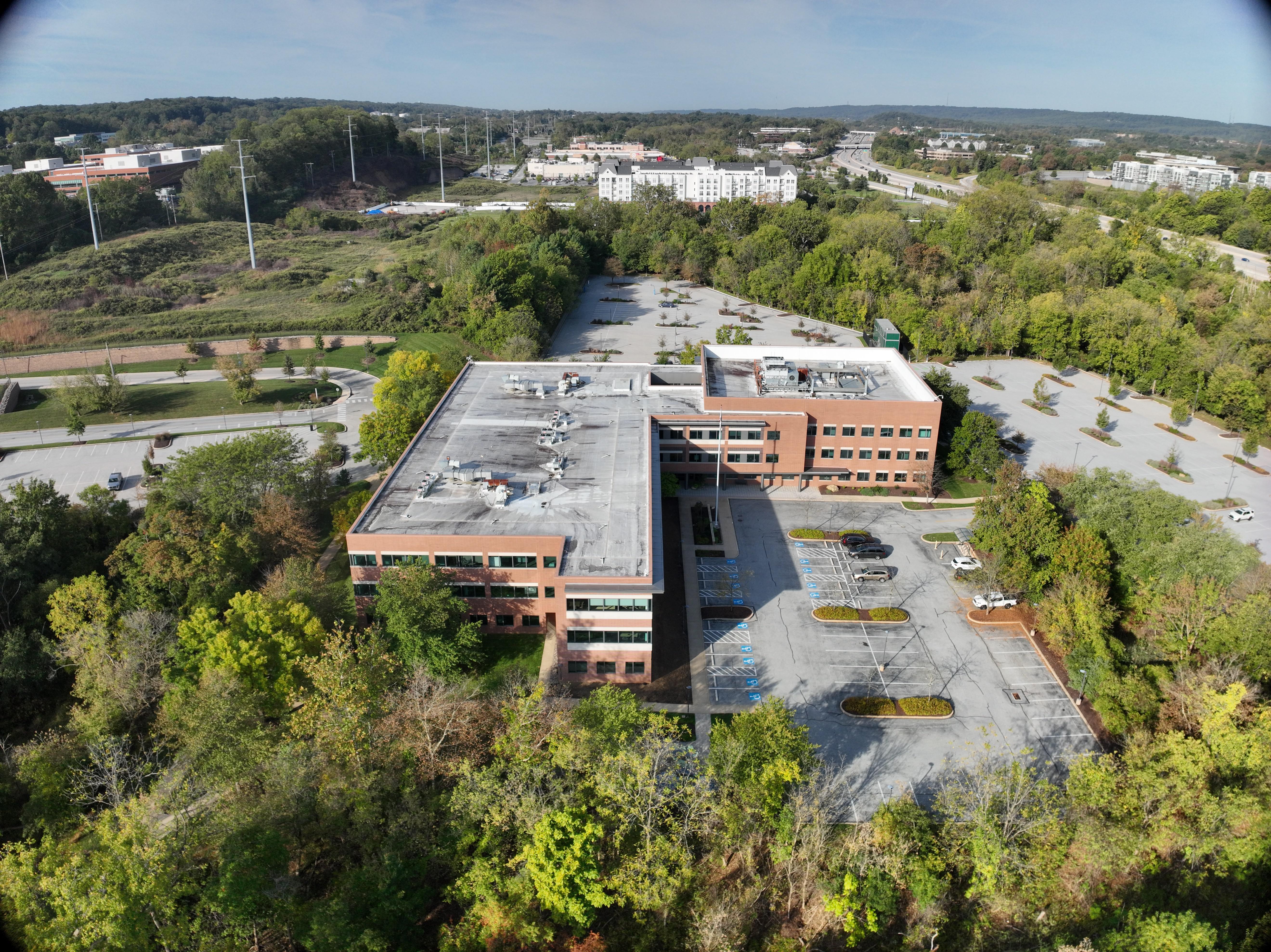 Aerial drone view of commercial office building complex with brick facade surrounded by lush green trees and parking facilities showcasing professional drone real estate and commercial property photography